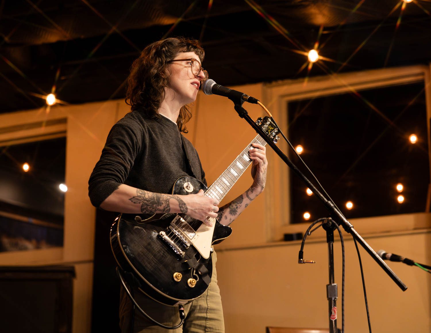 A solo musician performing on stage at a coffee shop, holding an electric guitar and singing into a microphone under ambient lighting.
