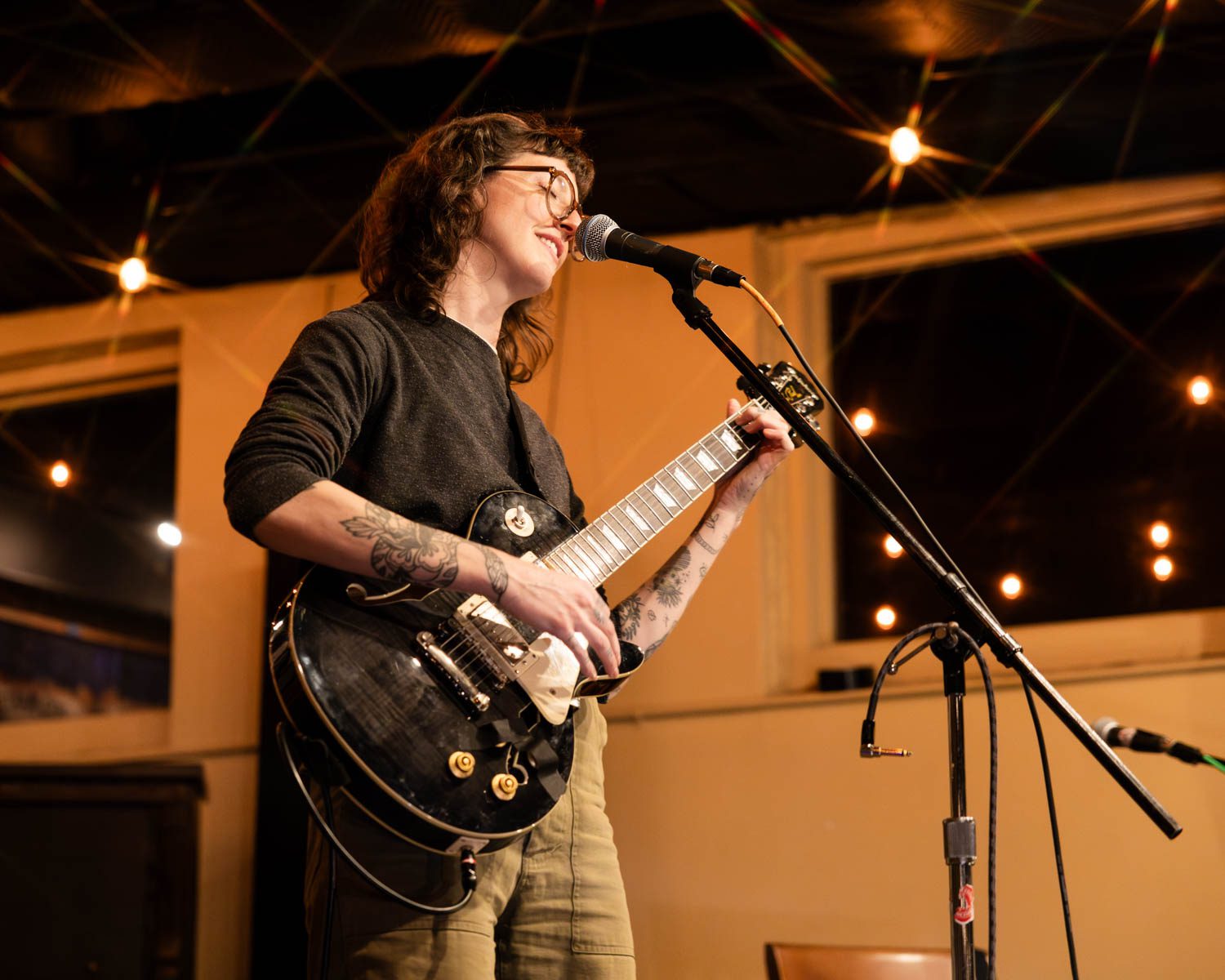 A woman performing on stage with a guitar, wearing glasses and a black sweater, smiling as she sings into a microphone at a cozy venue with warm lighting.
