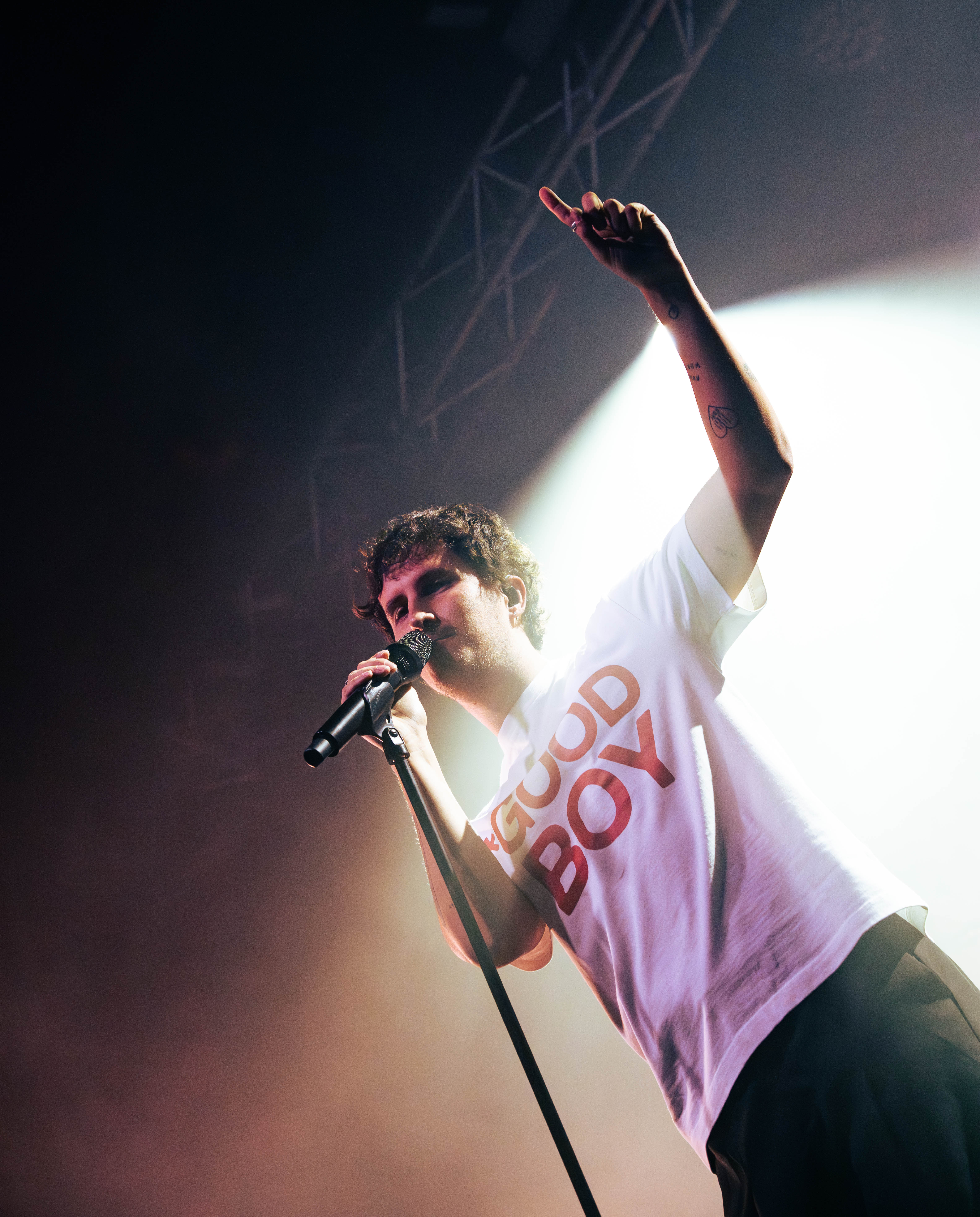 A performer singing into a microphone on stage during a live concert, wearing a white t-shirt with 'GOOD BOY' printed on it, illuminated by stage lights.