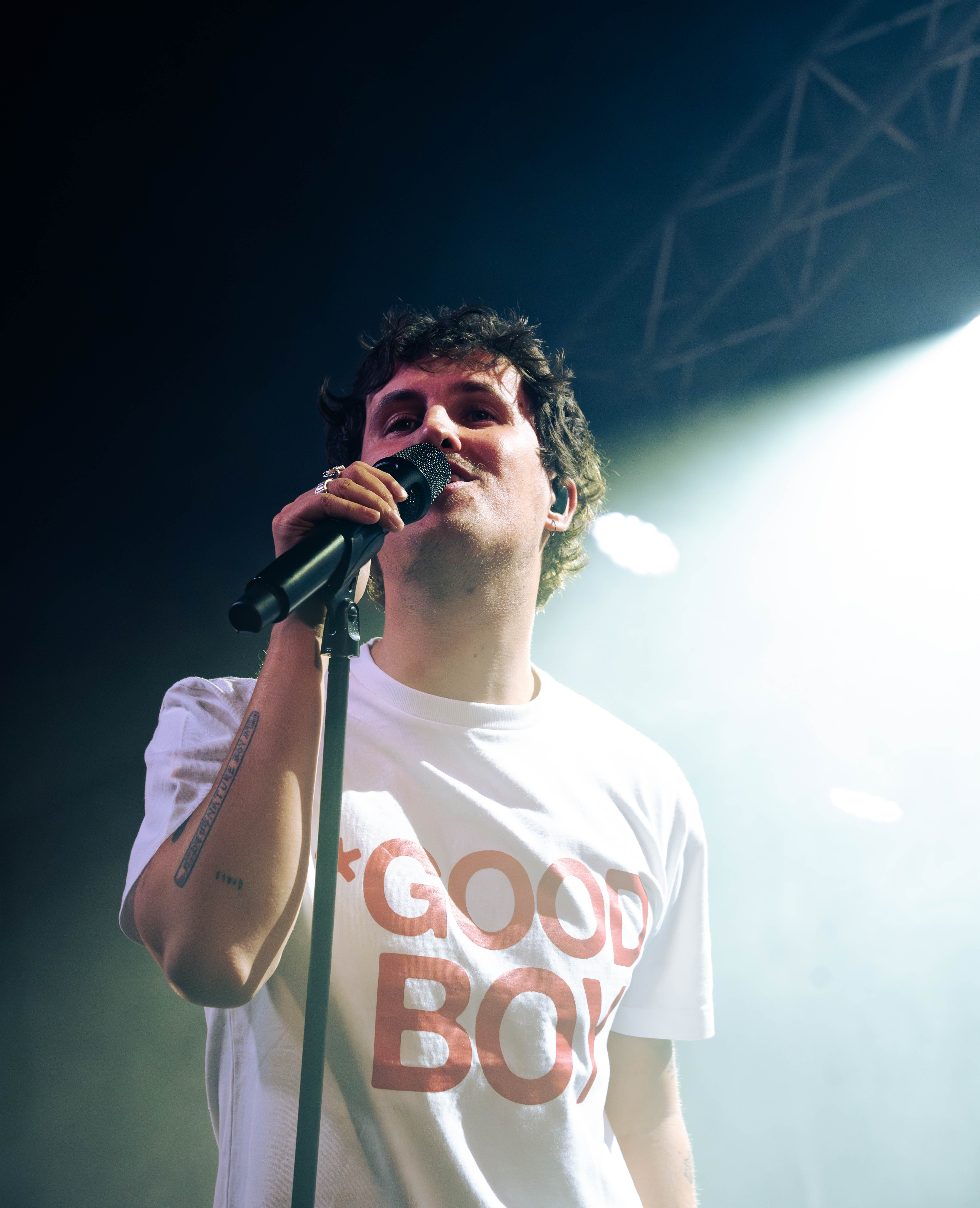 A performer in a white t-shirt with red text that reads '*GOOD BOY*', holding a microphone and singing on stage with bright stage lights behind him.