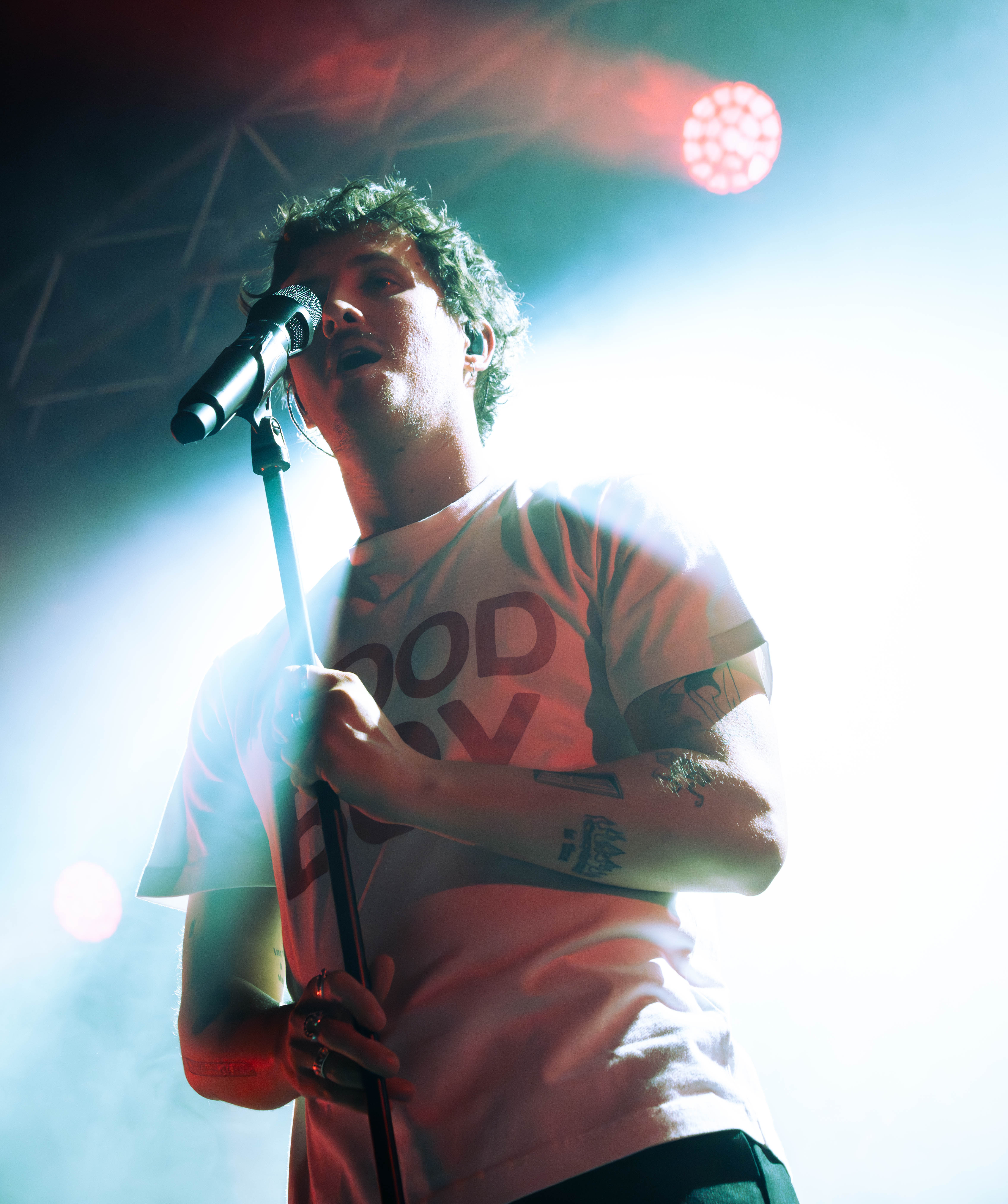 A male singer holds a microphone while performing on stage, illuminated by colorful stage lights.