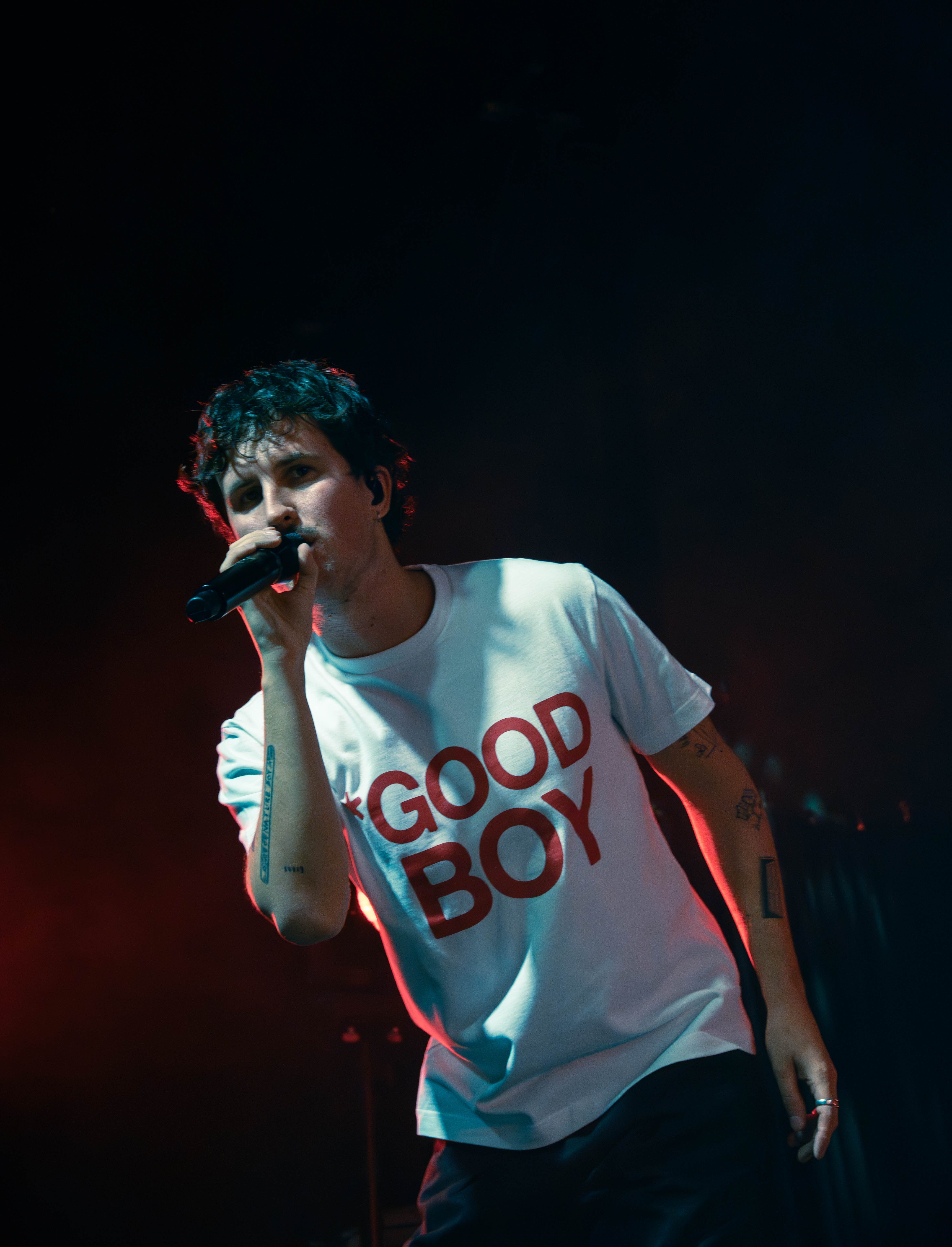 A performer singing into a microphone while wearing a white t-shirt with red text reading 'GOOD BOY', set against a dark background with colorful stage lights.