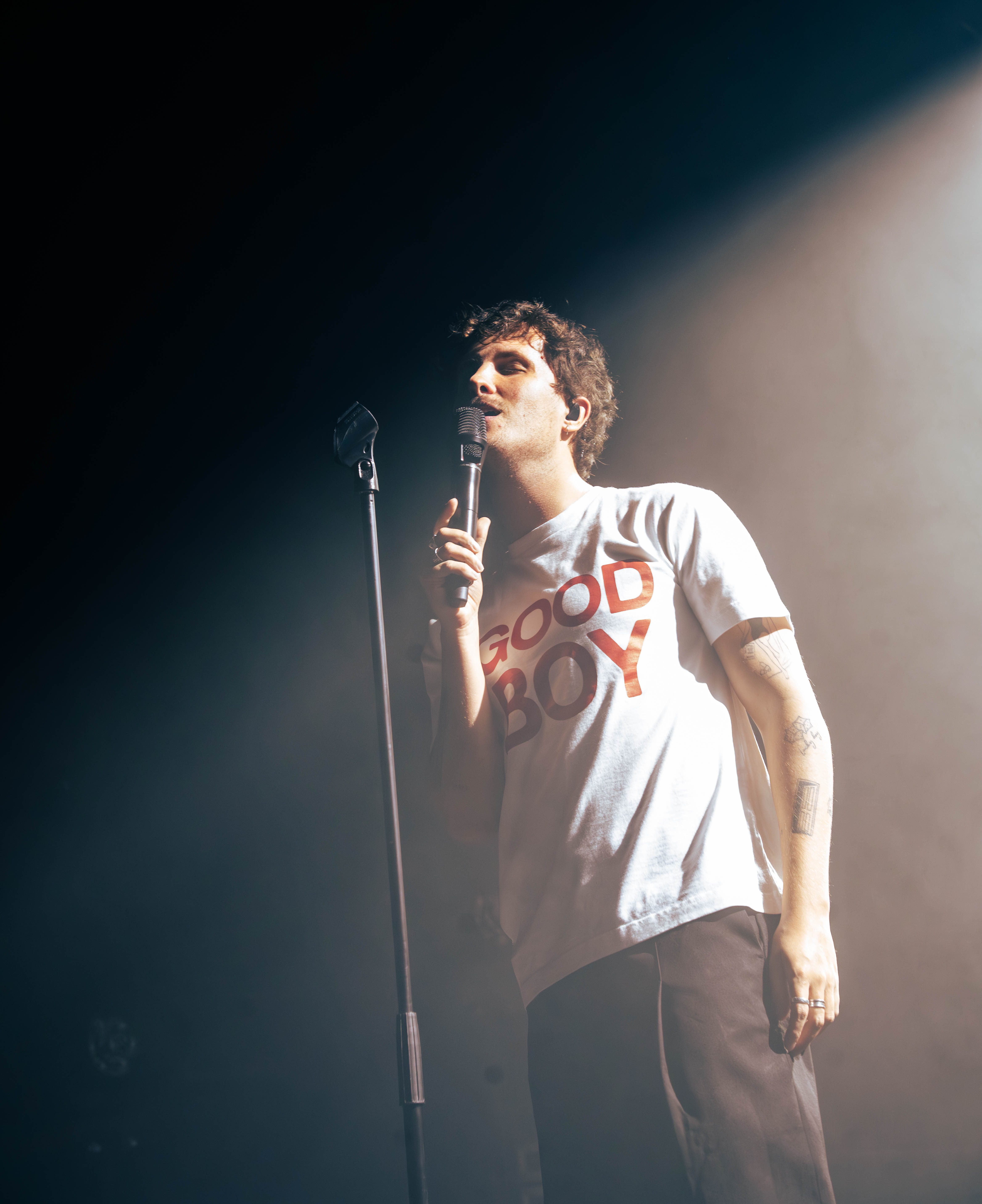 A musician performing on stage with a microphone, wearing a white t-shirt with 'GOOD BOY' printed in red, surrounded by dim lighting and smoke effects.