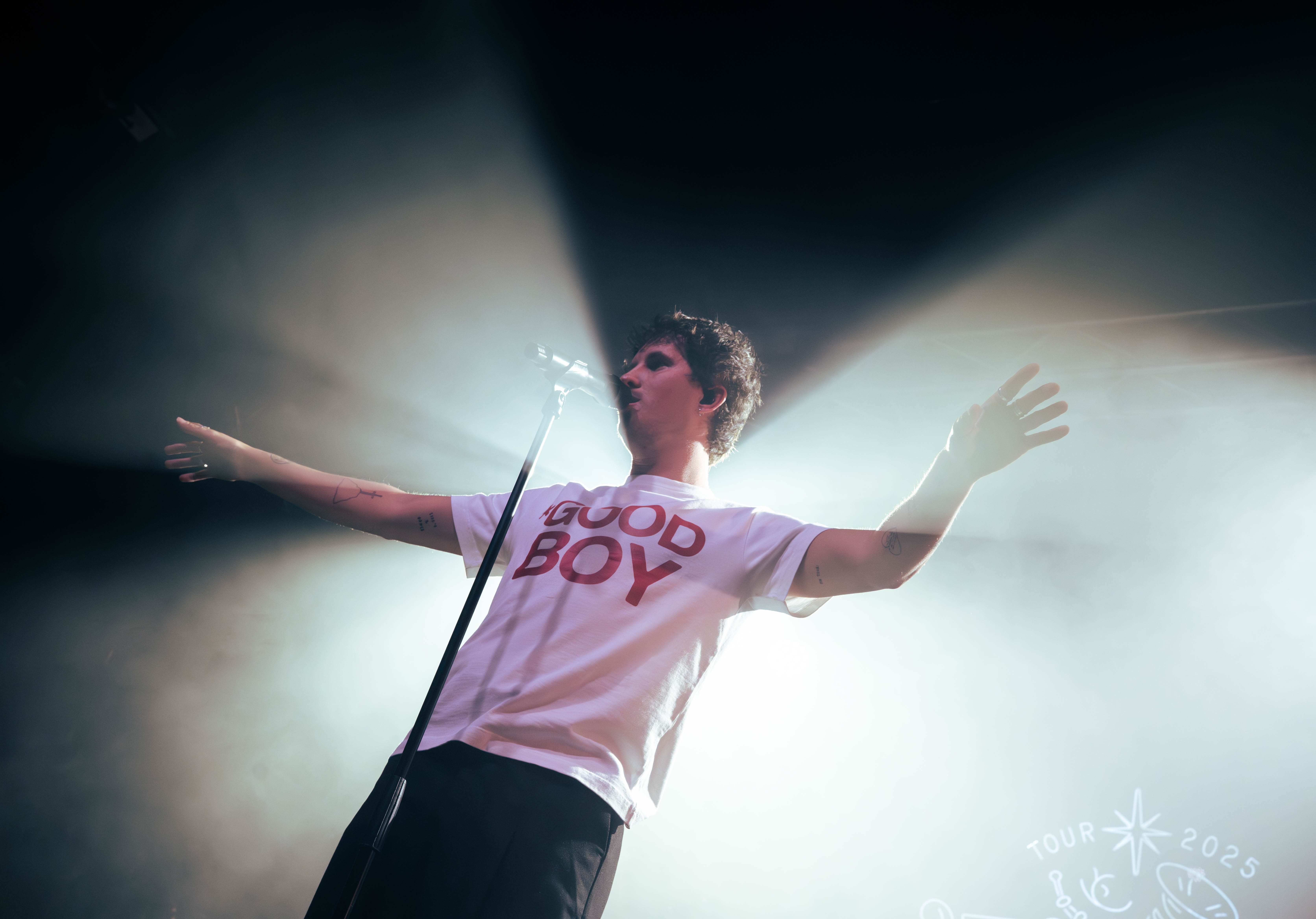 A performer stands on stage with arms outstretched, wearing a white t-shirt with the text 'GOOD BOY' in red. The background features dramatic lighting effects and a microphone stand.