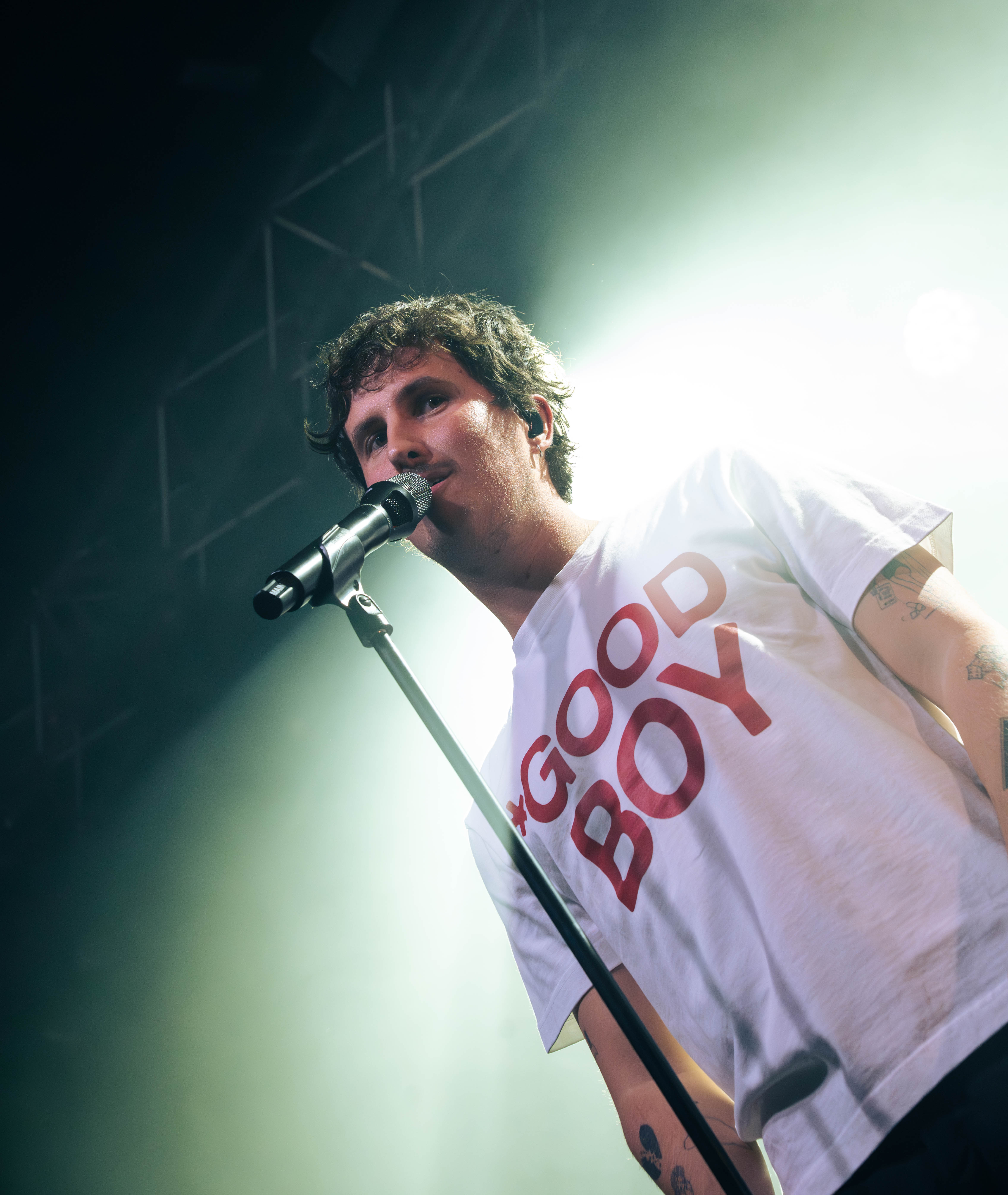 A musician performing on stage, wearing a white t-shirt with 'GOOD BOY' printed in red while holding a microphone.