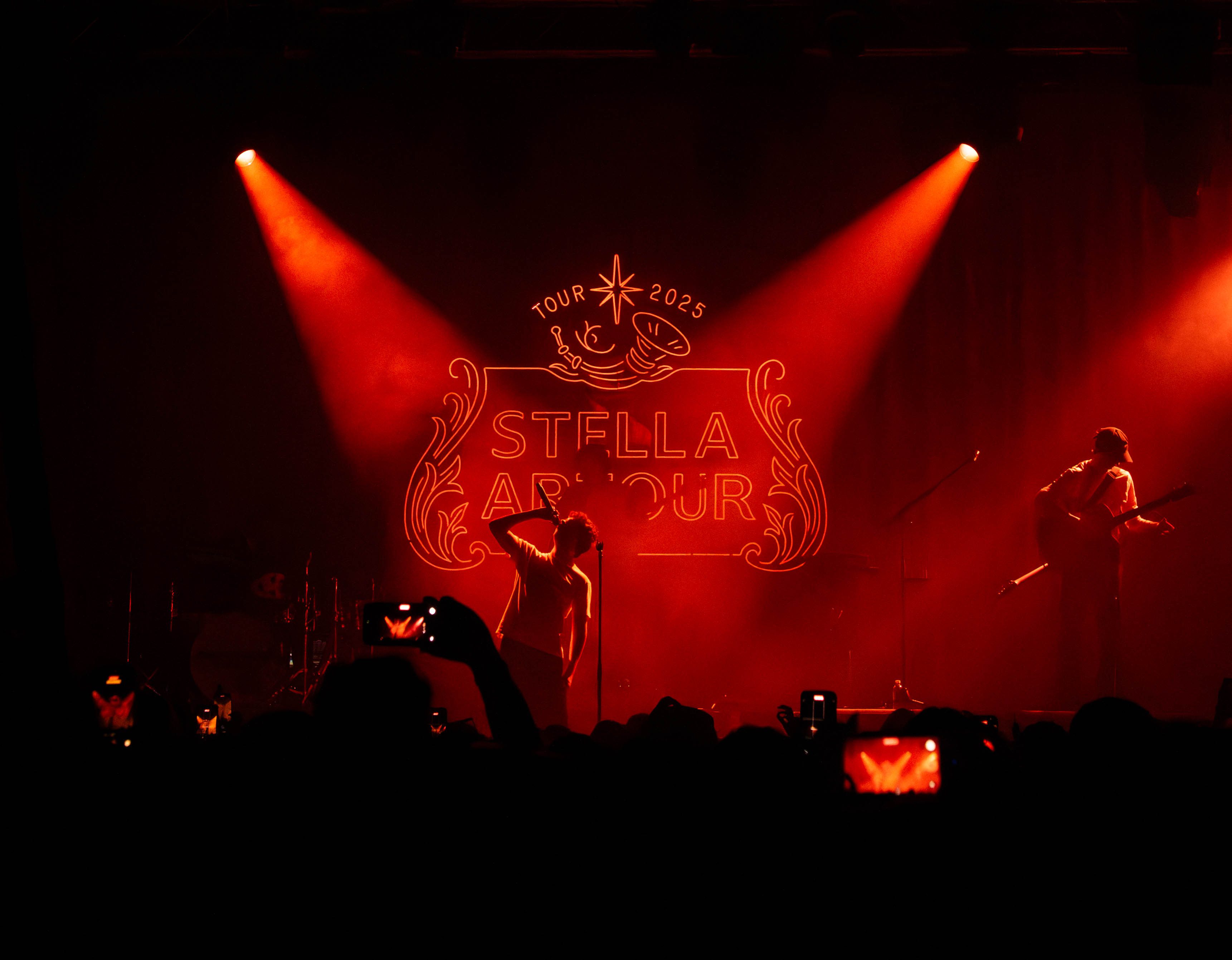 A performer on stage under dramatic red lighting, with a 'STELLA ARTOUR 2025' banner in the background. The crowd is visible in the foreground, capturing the moment with mobile devices.