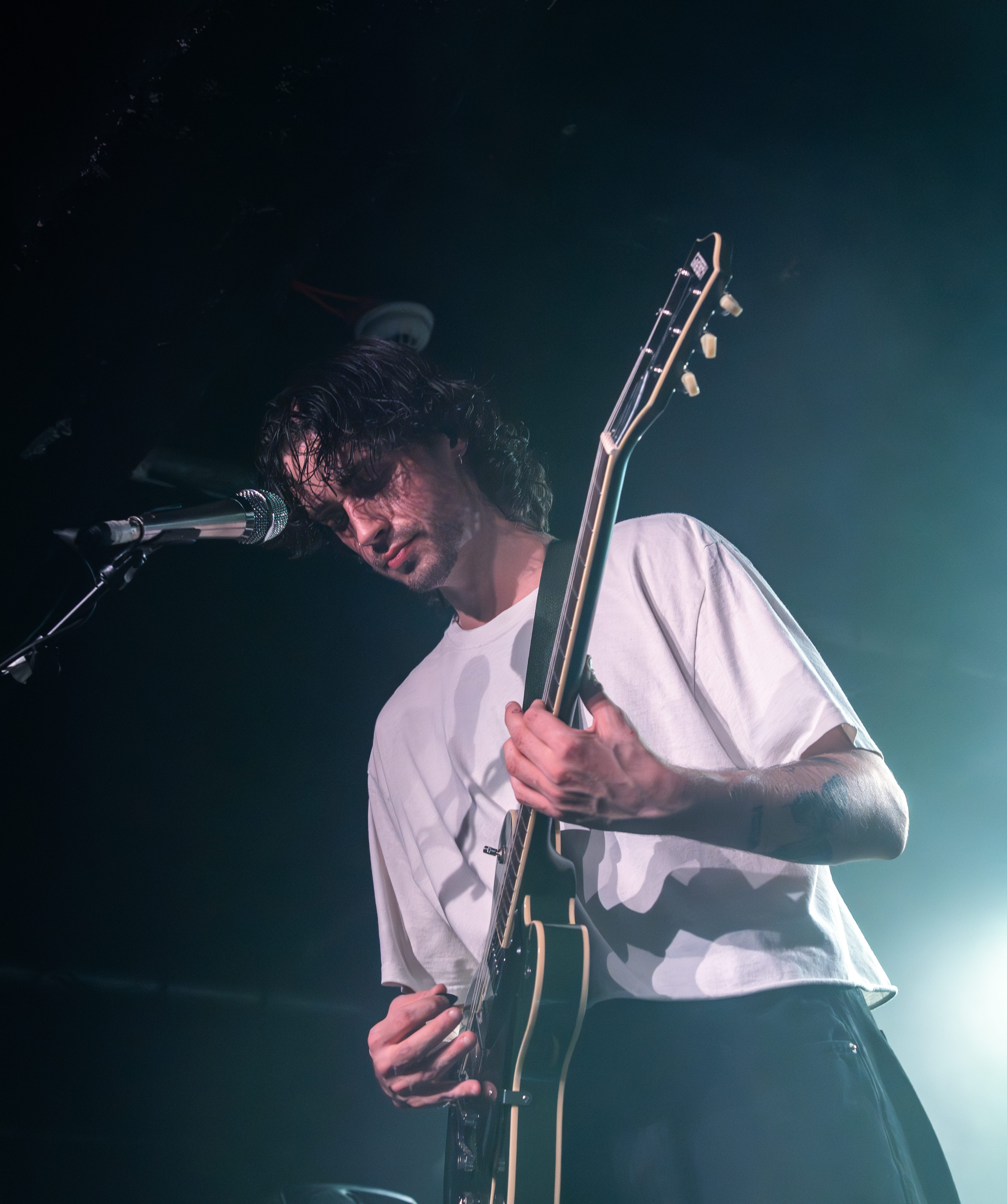 A musician performing on stage, playing an electric guitar with focused expression and a microphone nearby.