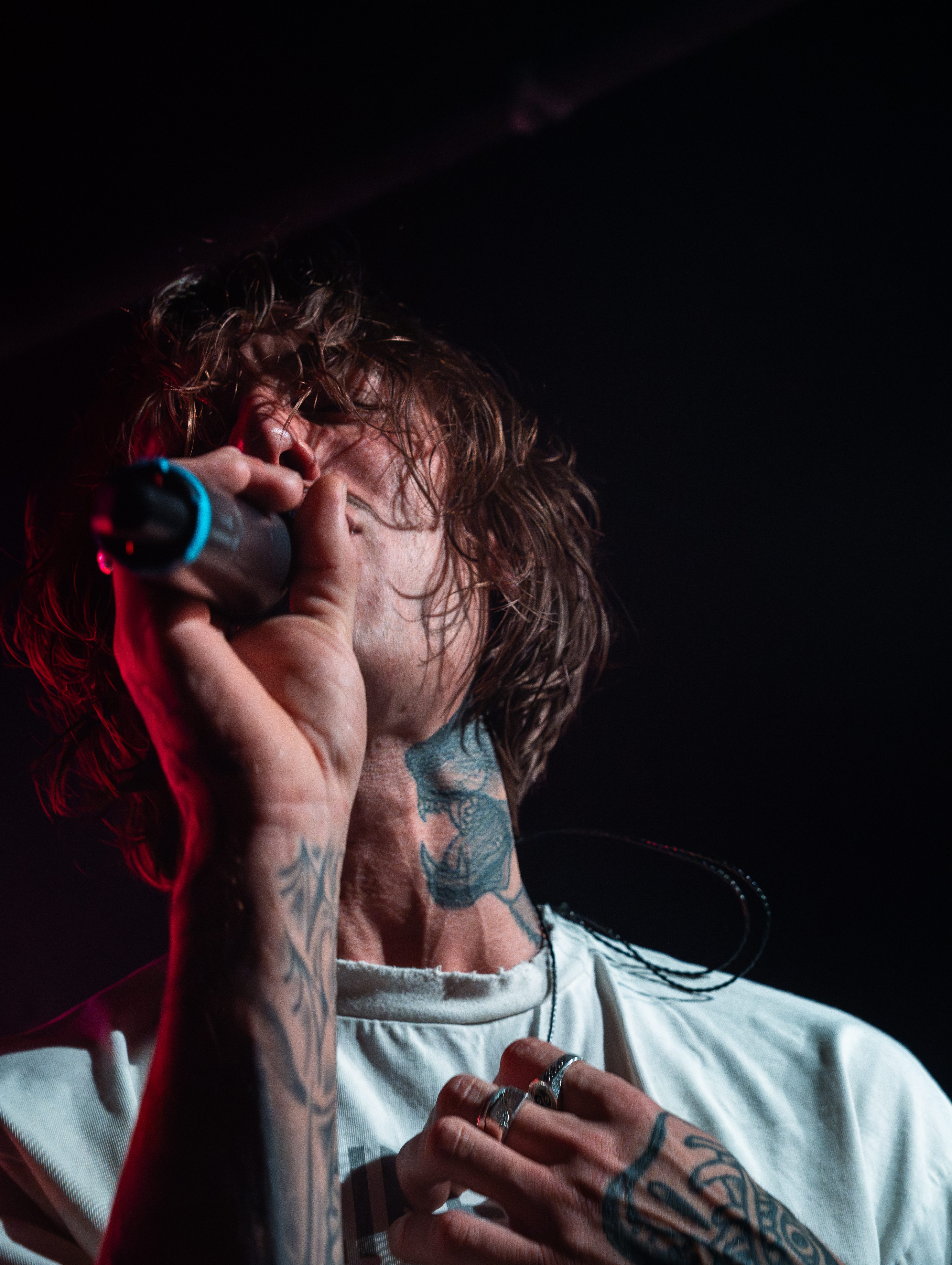 Close-up of a male singer passionately performing on stage, holding a microphone with tattoos visible on his arm and neck.