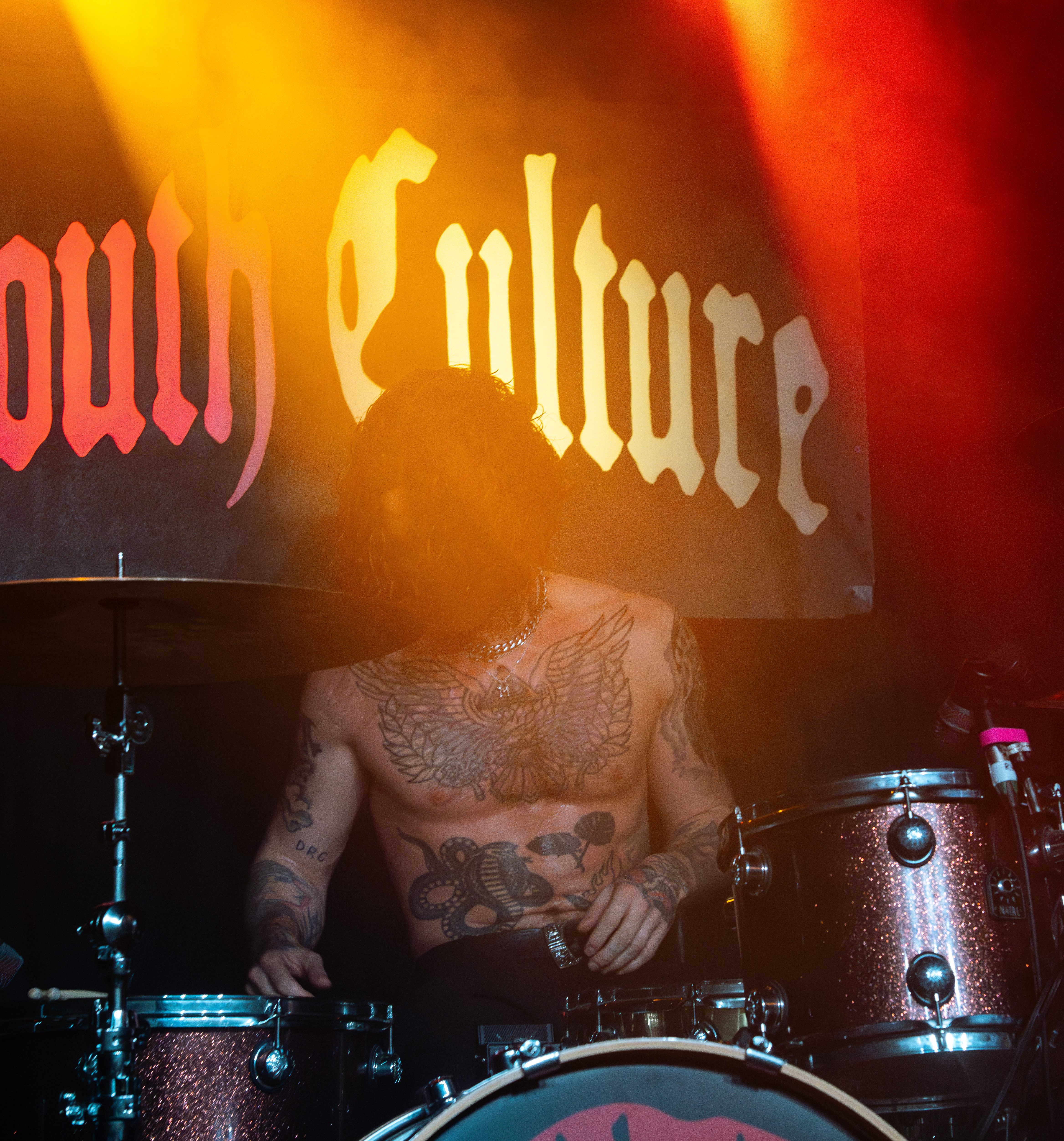 A drummer from the band Mouth Culture performing on stage, surrounded by colorful stage lights, with the band name displayed in the background.