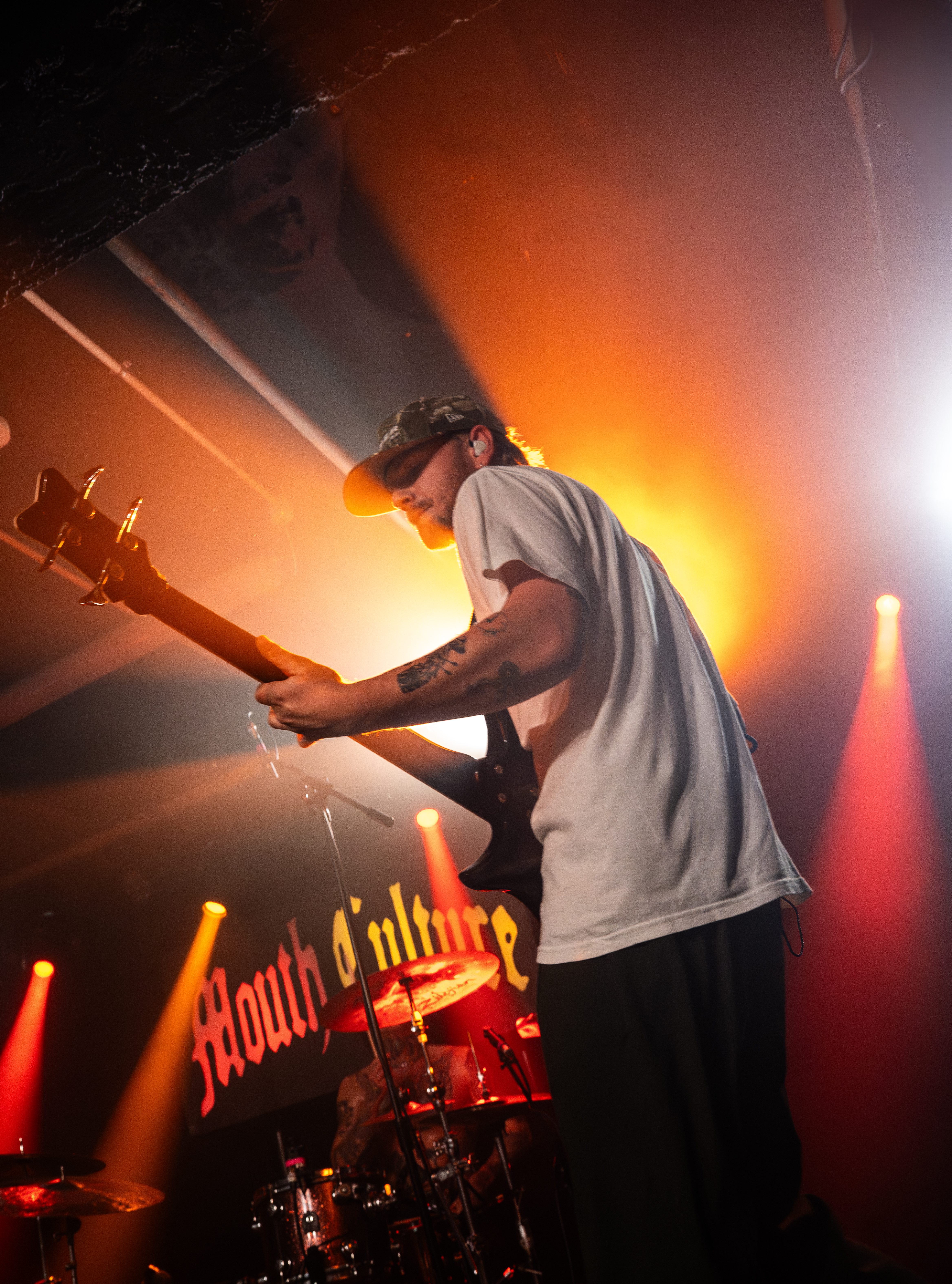A male guitarist performing on stage, wearing a baseball cap and a white t-shirt, with dramatic lighting in the background featuring the band name 'Mouth Culture'.