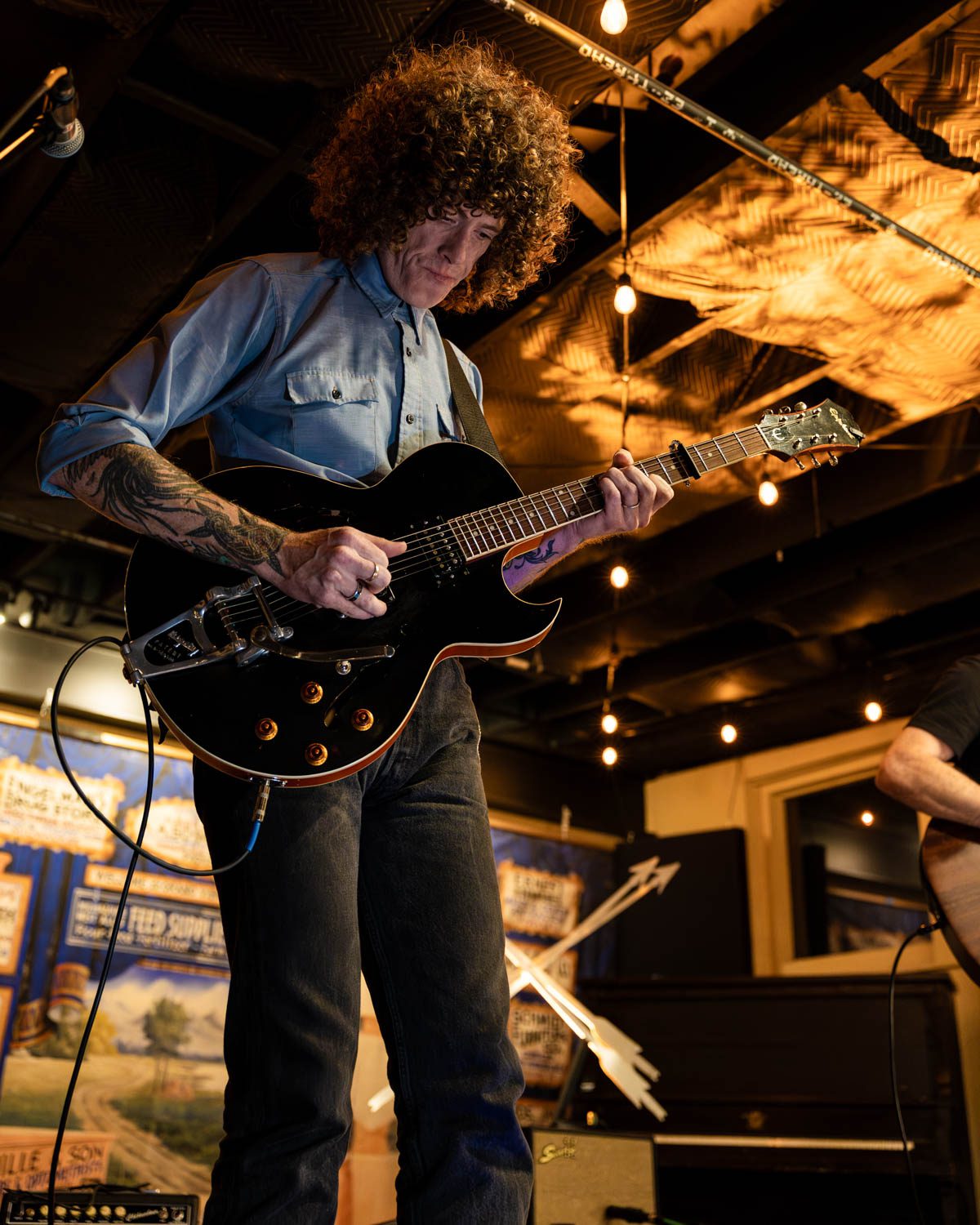 A musician with curly hair playing an electric guitar on stage, illuminated by warm string lights in a cozy venue.