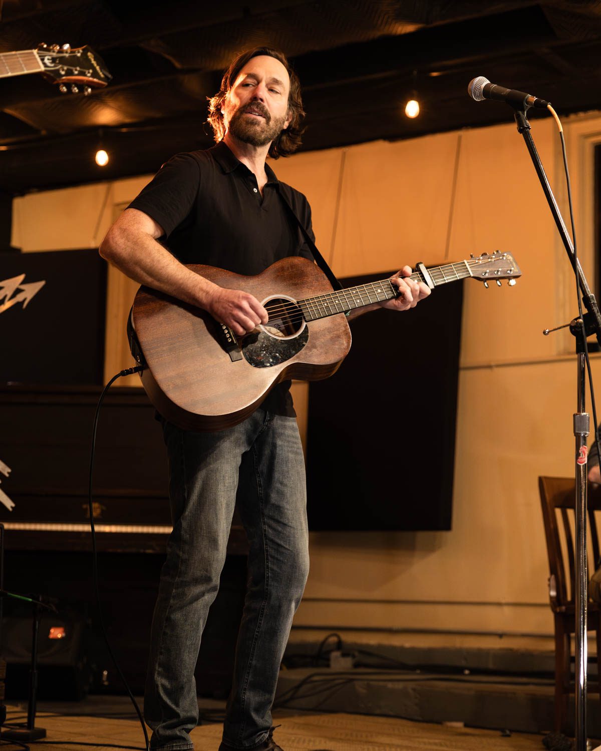 A musician performing on stage with an acoustic guitar, singing into a microphone, in a cozy venue with warm lighting.