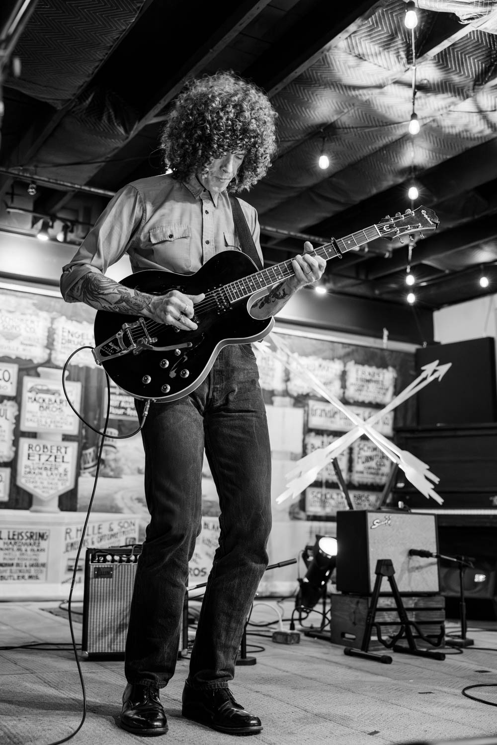 A musician with curly hair playing a black guitar on stage, dressed in a button-up shirt and jeans, with a relaxed atmosphere and string lights in the background.