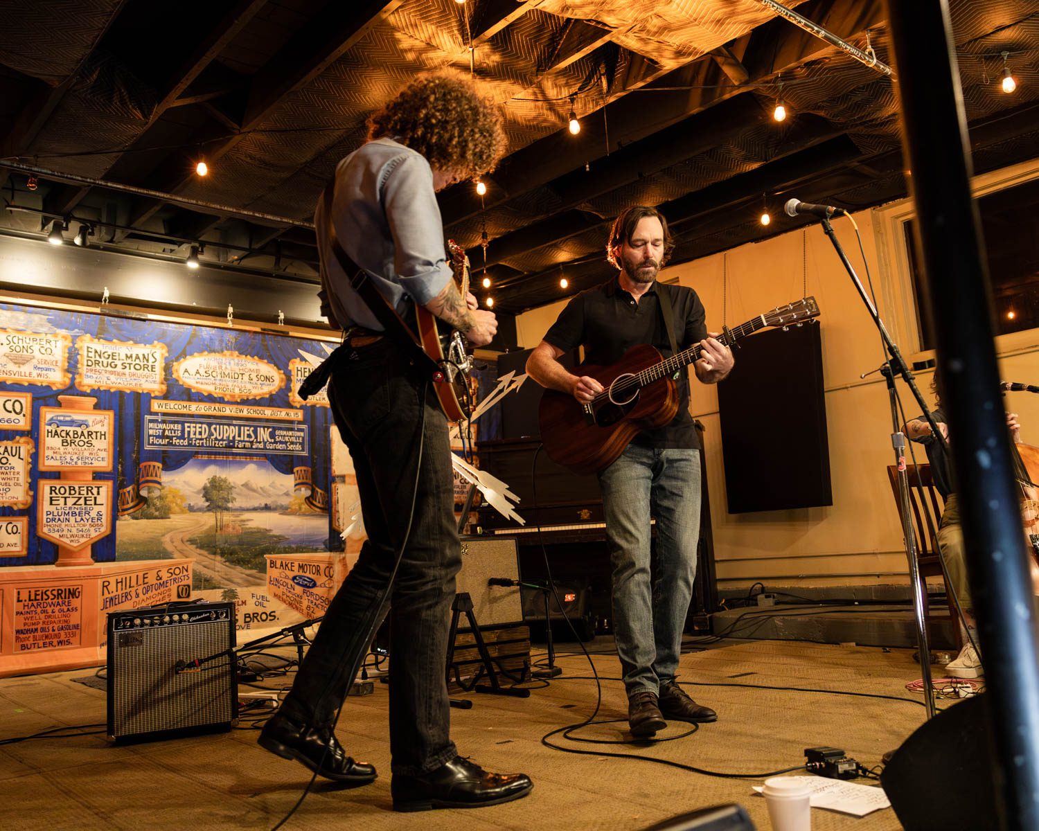 Two musicians performing on stage at Anodyne Coffee in Milwaukee, Wisconsin, with a colorful backdrop featuring vintage advertisements.