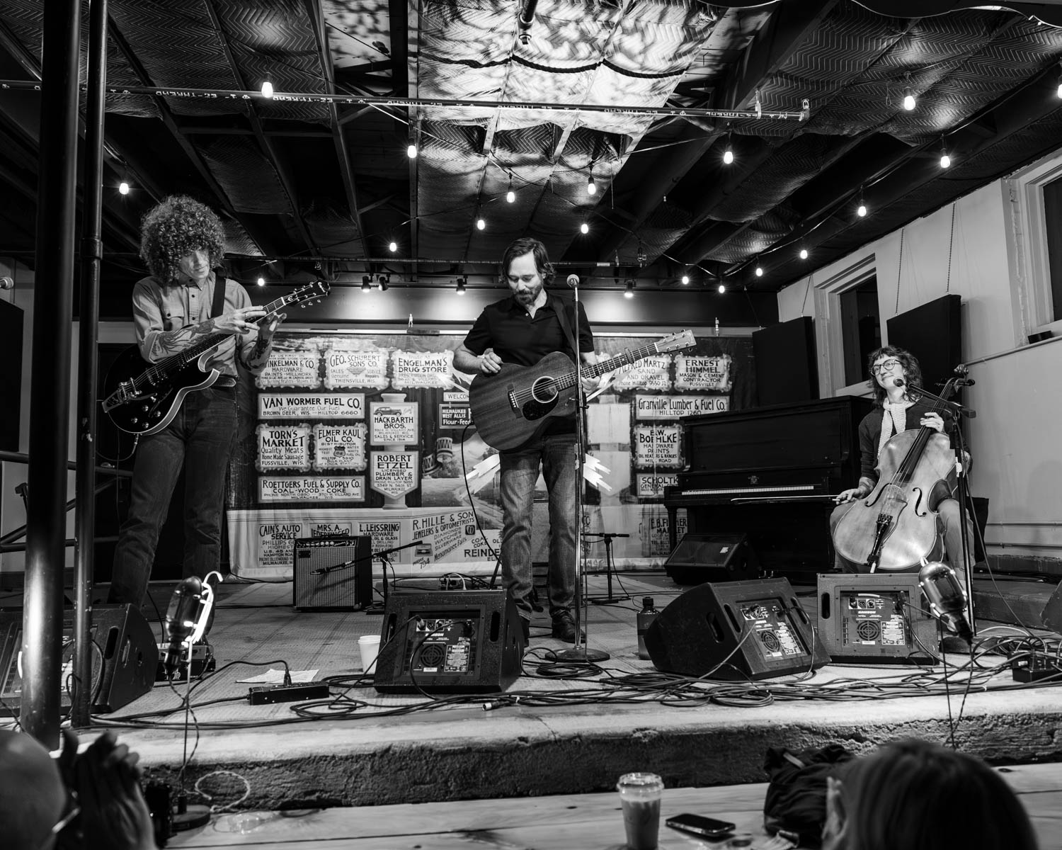 A black and white photograph of the band Matt Pond PA performing live at Anodyne Coffee in Milwaukee, Wisconsin. The stage features three musicians: a guitarist on the left, Matt Pond playing an acoustic guitar in the center, and a cellist on the right. The background includes vintage signs and string lights above, creating an intimate music venue atmosphere.