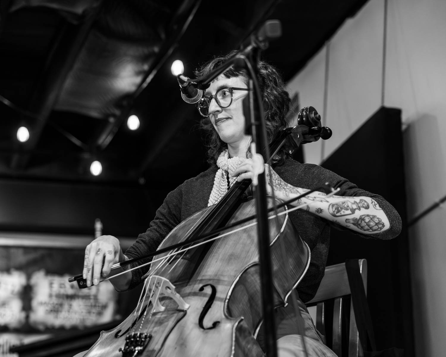 A woman playing the cello while sitting on a chair, with a microphone in front of her. She has curly hair and is wearing glasses. The background shows low lighting with hanging lights.