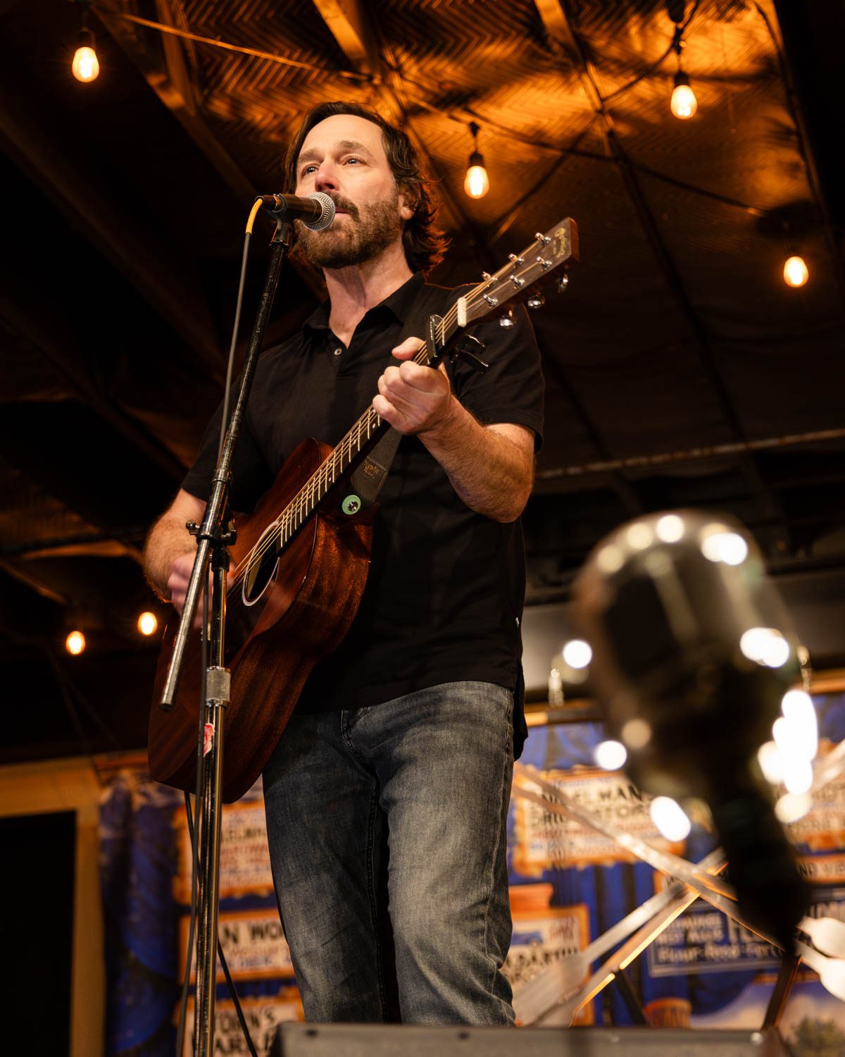 A male musician performing on stage with an acoustic guitar, microphone in front, and warm ambient lighting.