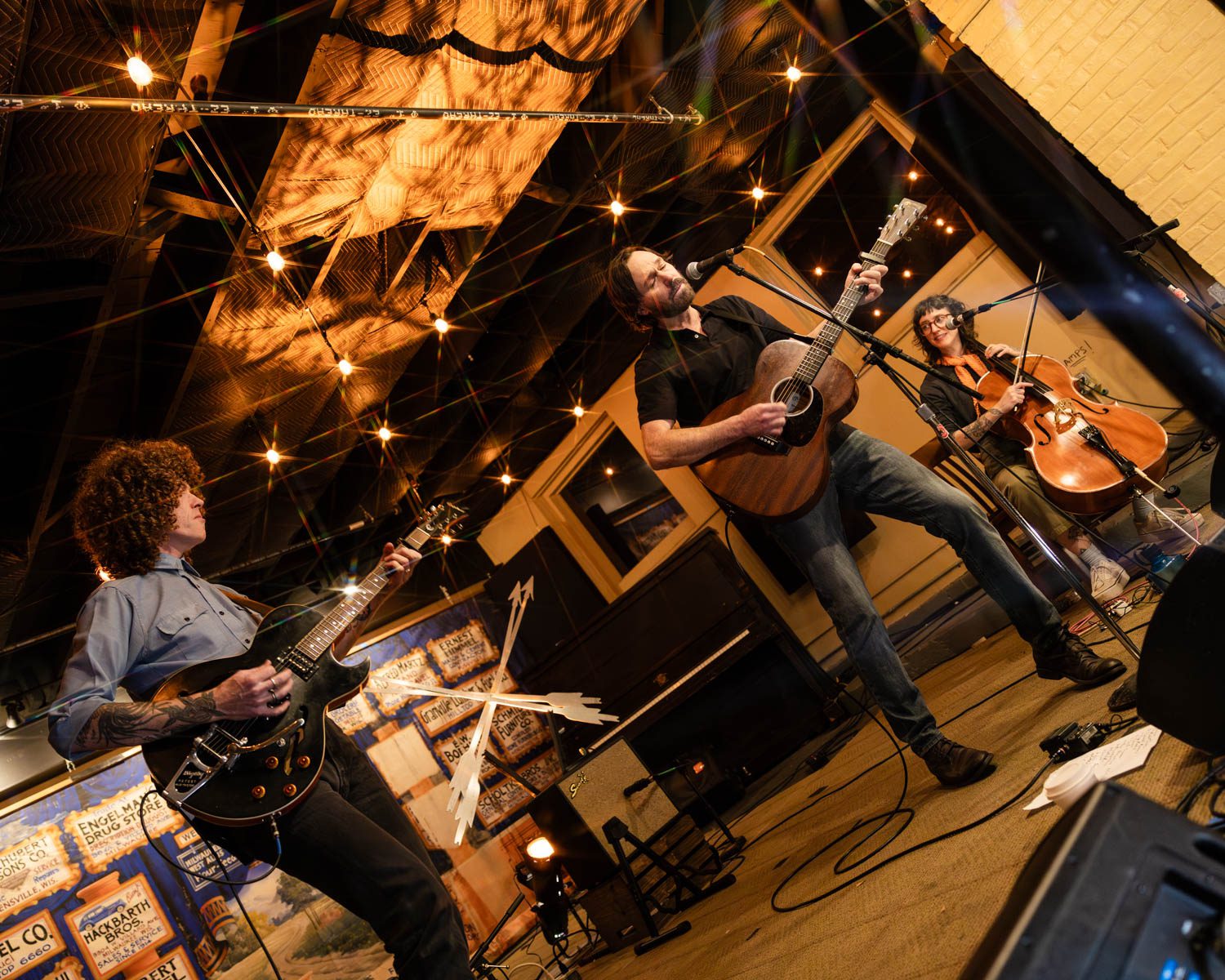A band performing on stage in a cozy coffee shop, with a guitarist, a vocalist playing an acoustic guitar, and a cellist, all under warm string lights.