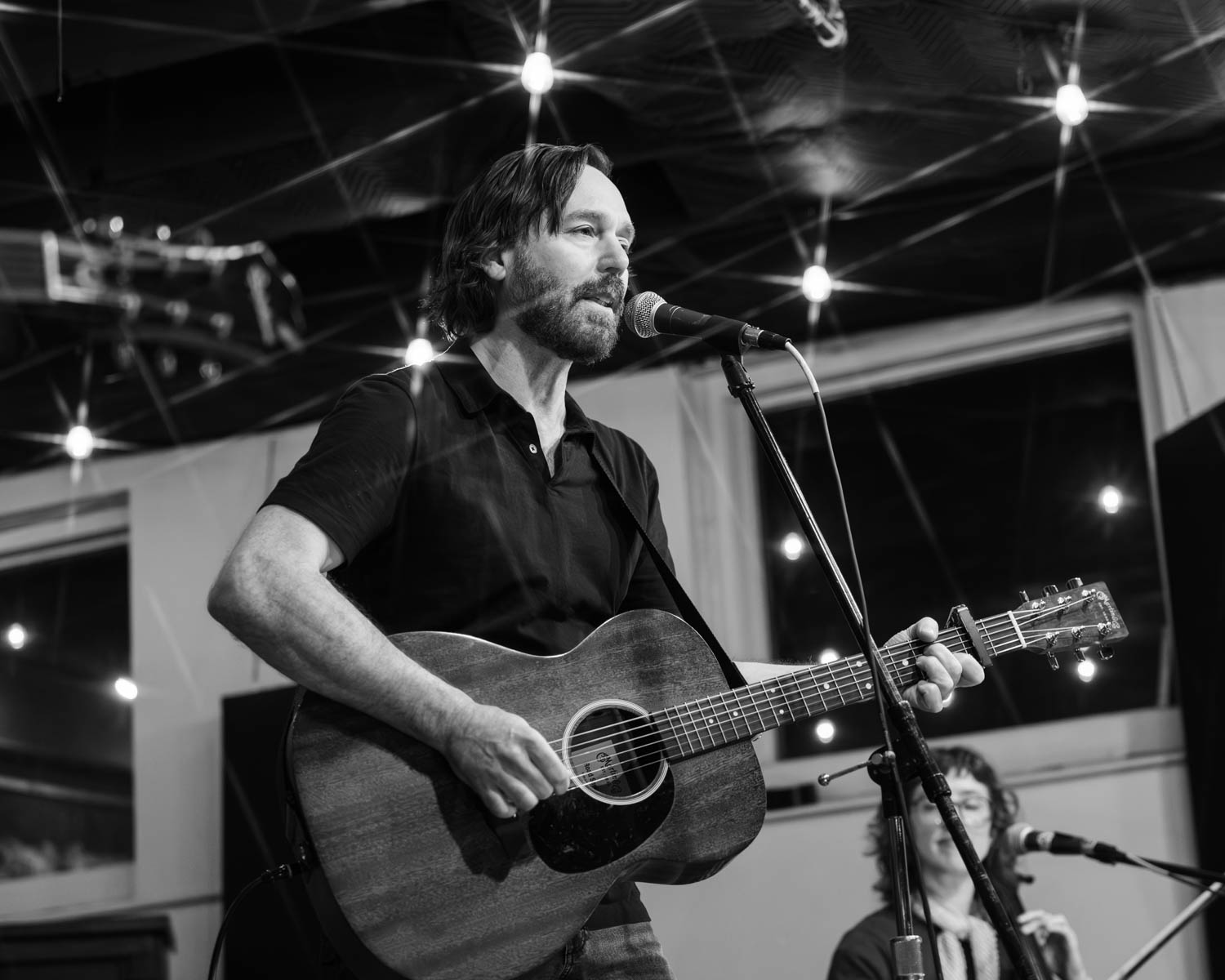 A man with a beard plays guitar and sings into a microphone on stage, while a woman with a microphone stands behind him. The scene is illuminated with soft lights and a cozy atmosphere.