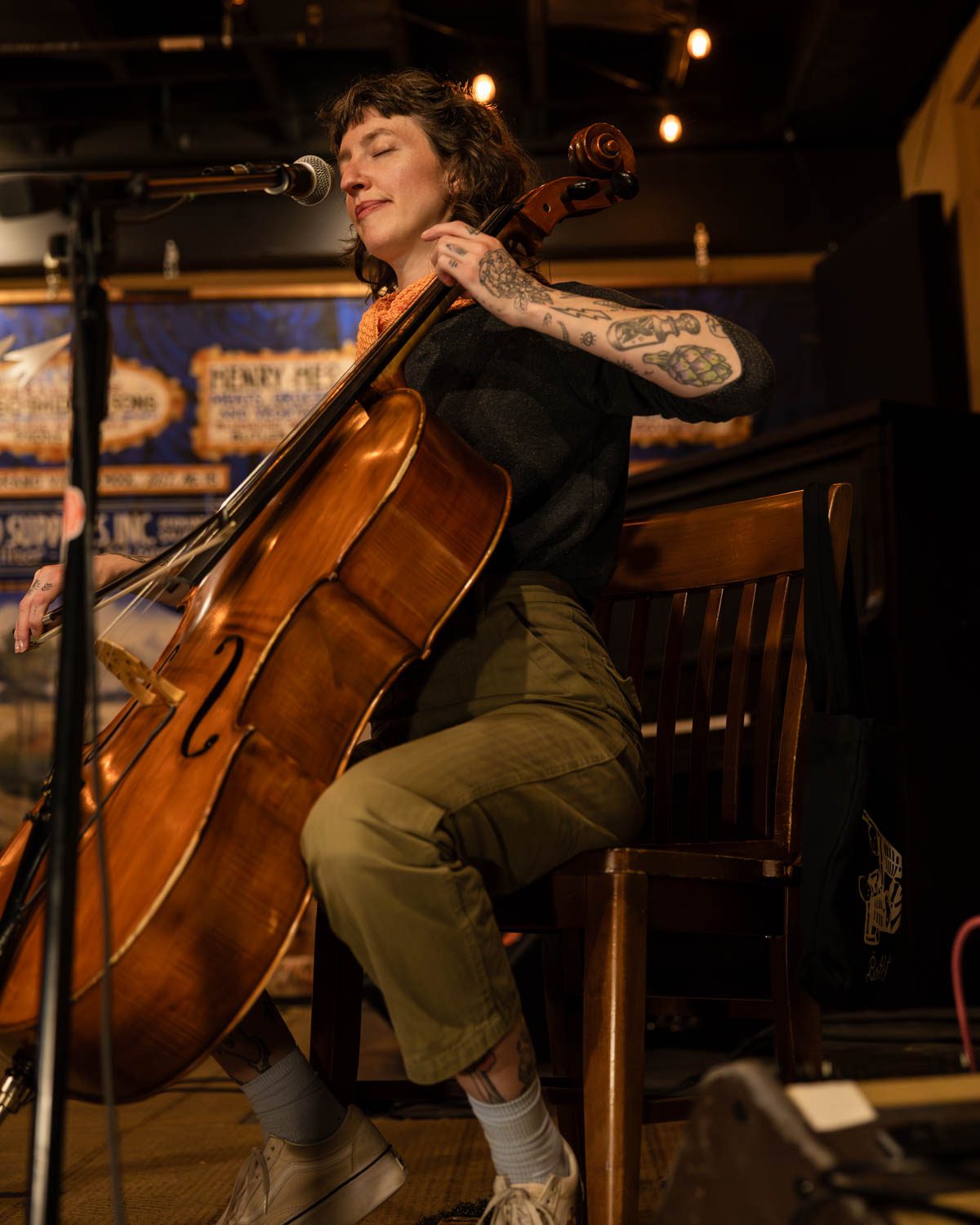A woman with tattoos playing a cello while sitting on a chair, singing into a microphone in a cozy venue with warm lighting.