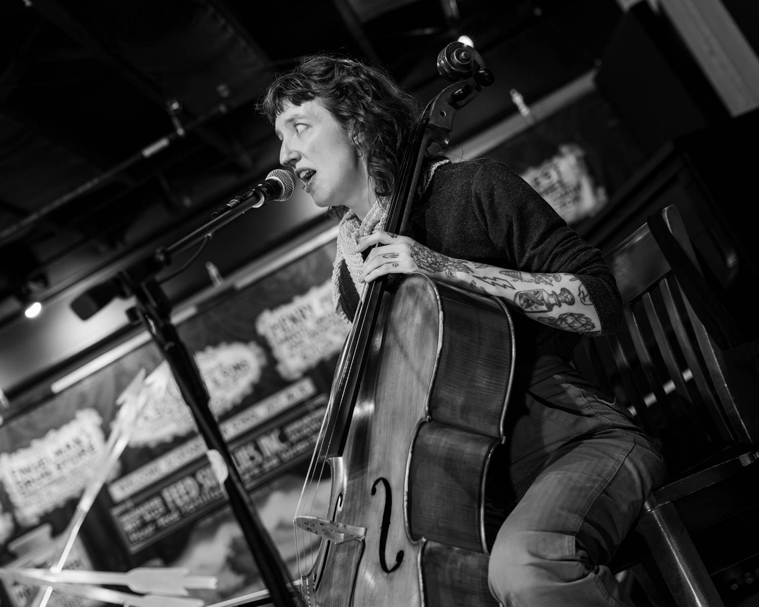 A black and white image of a woman singing into a microphone while playing a cello, with intricate tattoos visible on her arms. The background features performance posters.