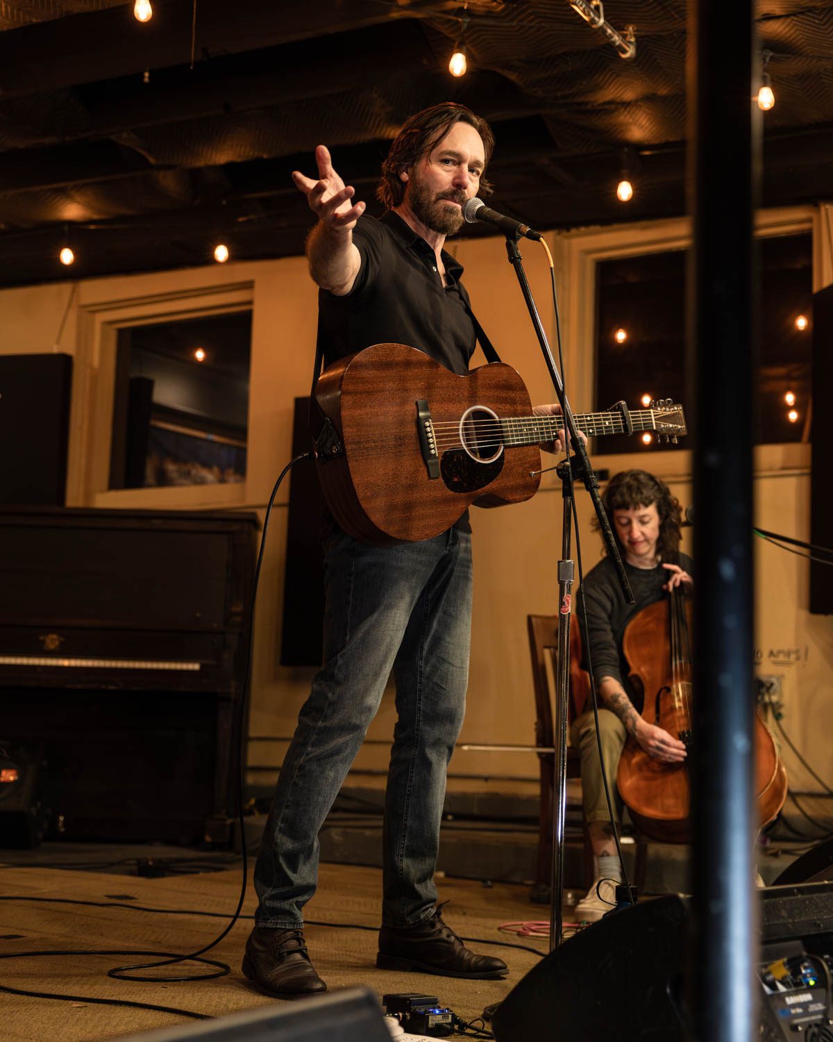 Musician performing with an acoustic guitar on stage, engaging the audience while a cellist accompanies in the background at a coffee shop concert.