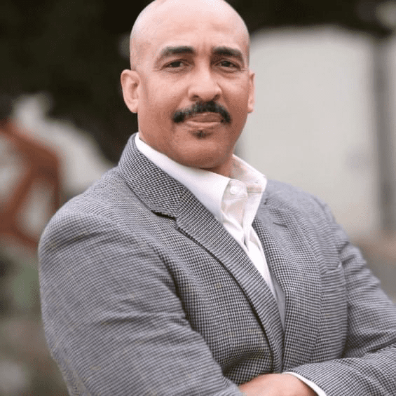 A confident man in a gray blazer poses with arms crossed in front of a blurred outdoor background.