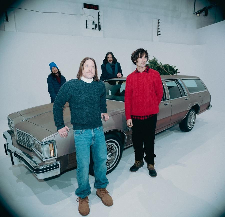 Four band members of The All-American Rejects posing next to a classic station wagon, decorated with a holiday tree, in a minimalist studio setting.