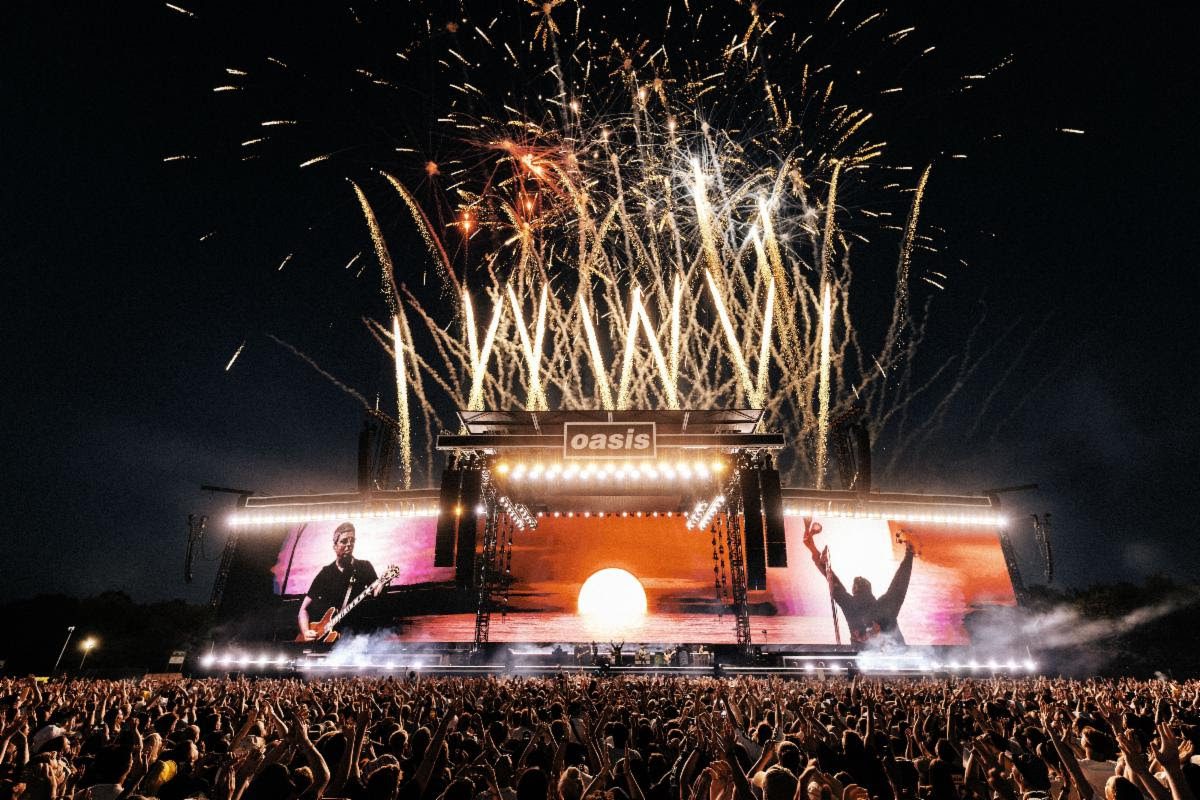 A large crowd at an Oasis concert with fireworks lighting up the night sky, showcasing a vibrant stage and a sunset backdrop.