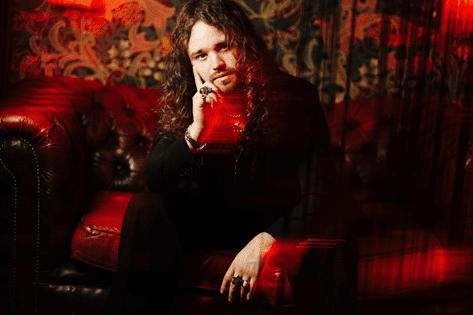 A male artist with long curly hair, wearing a black outfit, sits thoughtfully on a velvet couch with a colorful patterned background.