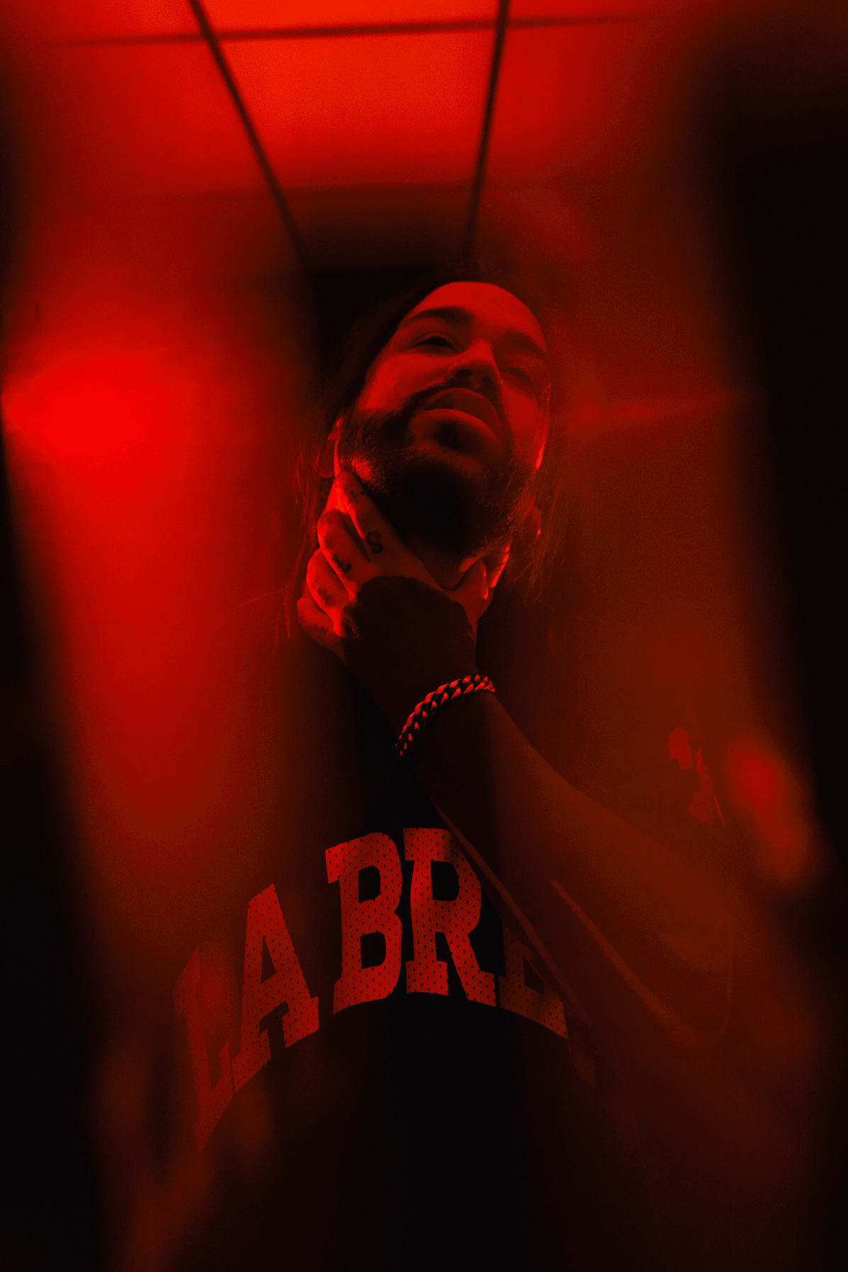 A moody, red-tinted photograph of a man with long hair and a beard, holding his neck thoughtfully, wearing a black shirt with 'LA BREA' written on it.